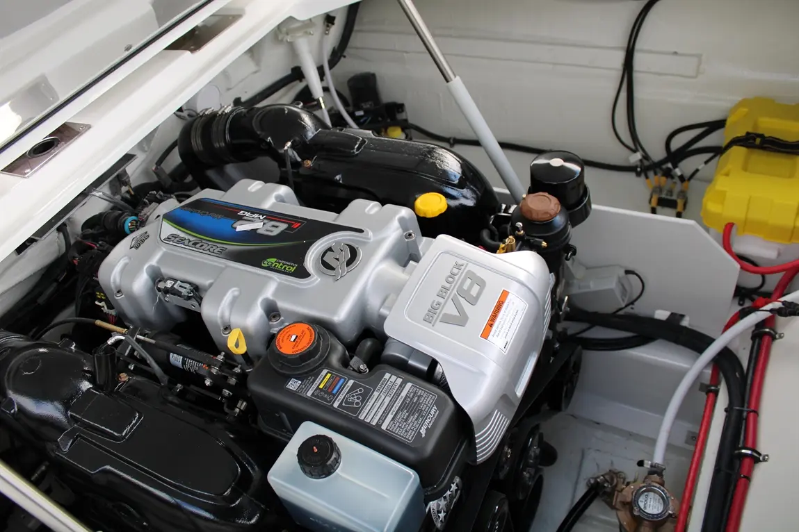 A big-block V8 engine viewed from above, featuring various hoses, caps, and a yellow reservoir in a boat engine compartment.